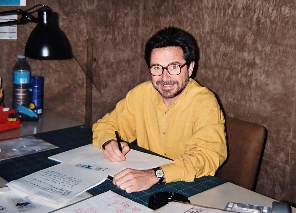 José Luis Gimeno con barba, gafas y camisa amarilla, sonriendo en su mesa de trabajo con un flexo y bocetos